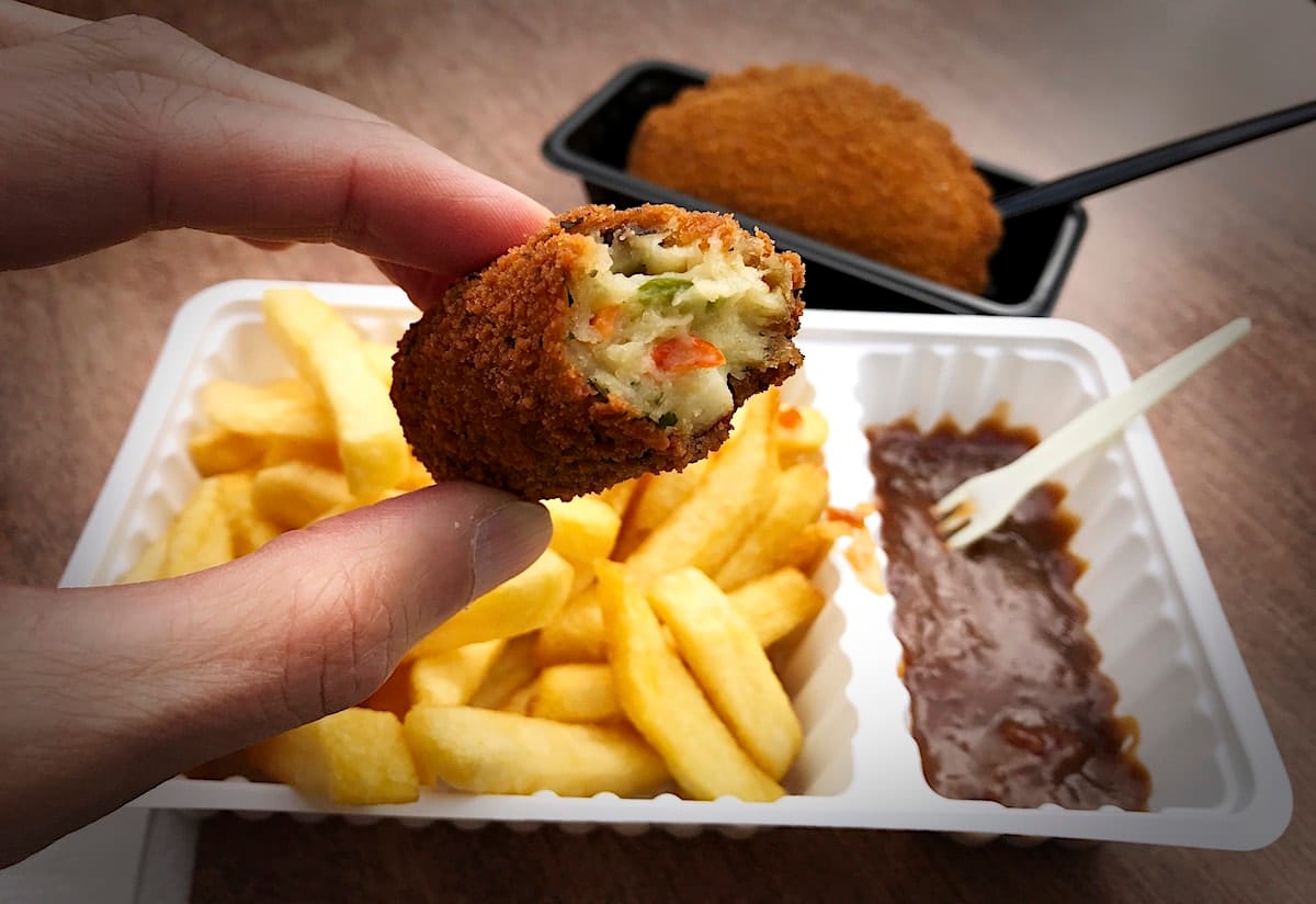 Eine angebissene Gemüsekrokette, dahinter Pommes mit Erdnuss-Sauce und ein anderer Snack, das Kaas-Soufflé